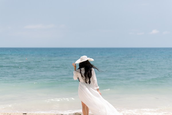 bride from behind walks towards sea on sandy beach wearing sun hat