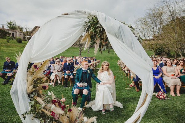 shot of bride and groom within the circular flowerbed frame guests behind them