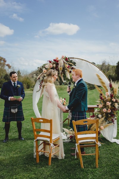 bride and groom in front of circular flower bed at alter to wedding they hold hands reading vows