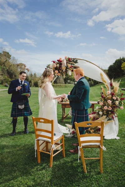 bride and groom in front of circular flower bed at alter to wedding they hold hands reading vows