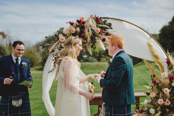 bride and groom in front of circular flower bed at alter to wedding they hold hands reading vows