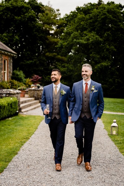 grooms walk in the courtyard smiling one is holding glass of champagne garden setting