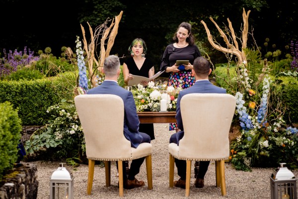 grooms sit in white chairs at alter setting with celebrant giving speech