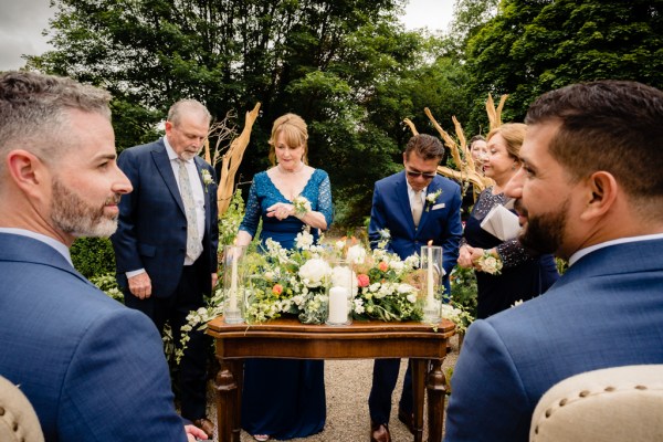 grooms sit in white chairs at alter setting with celebrant giving speech witnesses