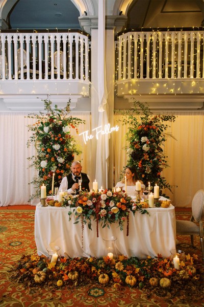 Bride and groom sit at table covered in flowers he's giving a speech