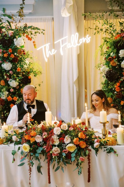 Bride and groom sit at table covered in flowers he's giving a speech