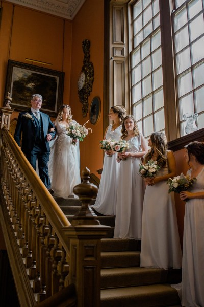 father of the bride walks down staircase bridesmaids stand on staircase in a row