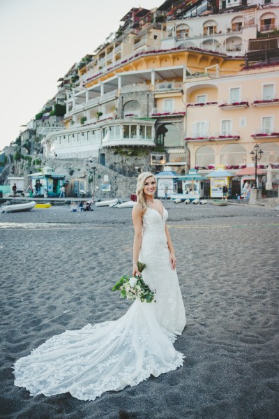 Bride walking on a beach holding her bouquet in front of buildings in Positano