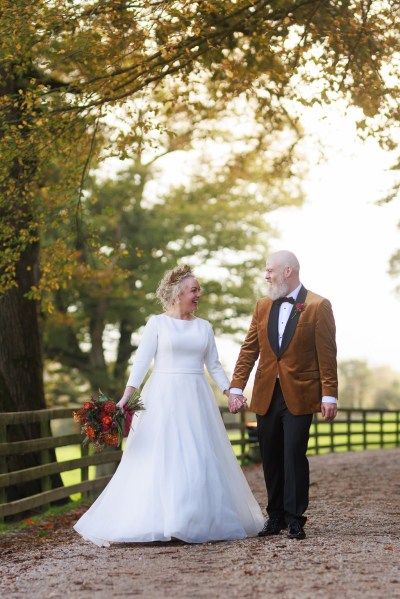 bride and groom stand in front of gate to forest garden setting trees surround they hold hands and walk