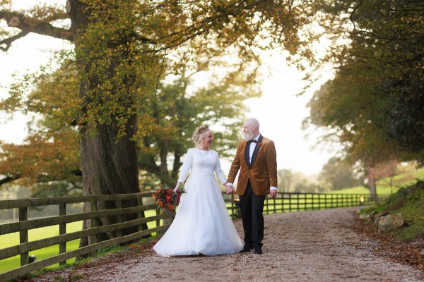 bride and groom stand in front of gate to forest garden setting trees surround they hold hands and walk