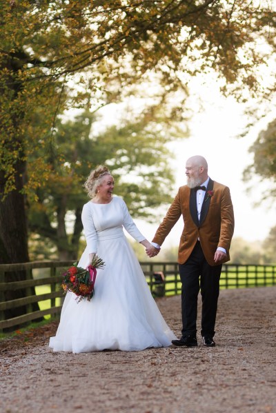 bride and groom stand in front of gate to forest garden setting trees surround they hold hands and walk