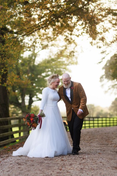 bride and groom stand in front of gate to forest garden setting trees surround they hold hands and walk and joke around