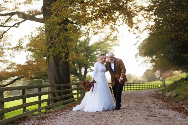 bride and groom stand in front of gate to forest garden setting trees surround they hold hands and walk and joke around