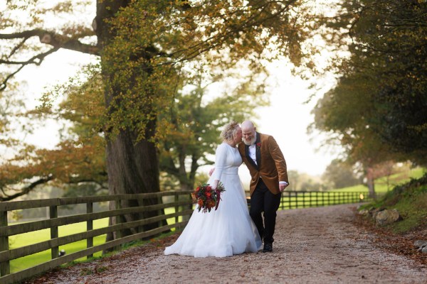 bride and groom stand in front of gate to forest garden setting trees surround they hold hands kiss