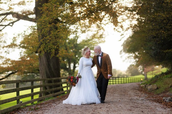 bride and groom stand in front of gate to forest garden setting trees surround they hold hands and walk and joke around