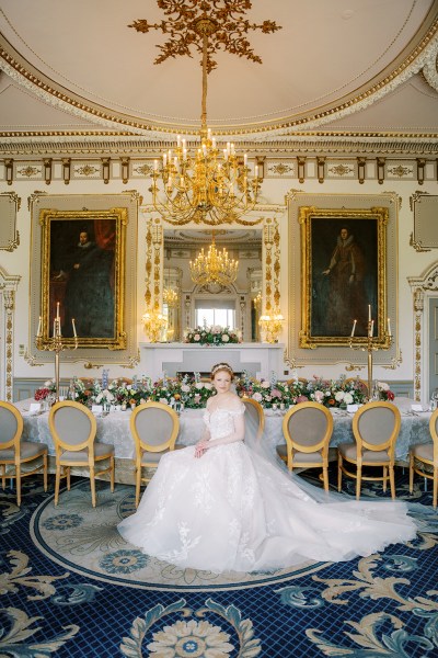 bride looks up hand over hand on lap shows off hair accessory dining room interior portraits on wall gold themed