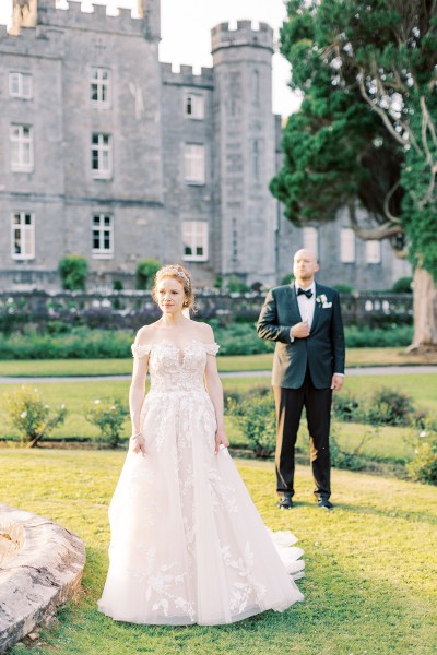 bride stands in front of groom on grass he's behind her waiting