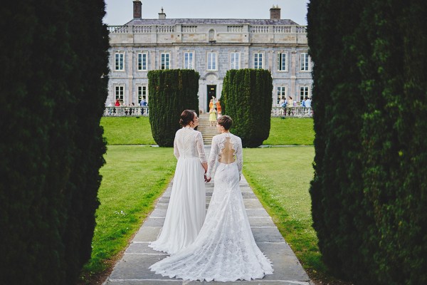 Brides walk from behind towards wedding venue along pathway holding hands