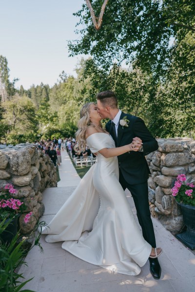 bride and groom kiss beside bricked wall pink flowers and trees surround