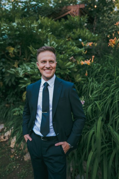 groom is all smiles as he stands in front of shrub in forest garden