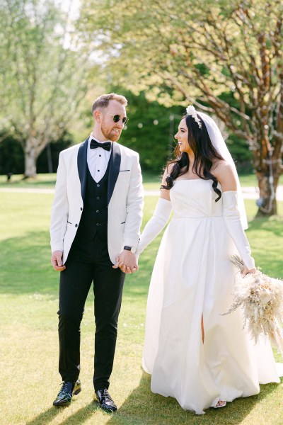 Bride and groom face each other standing on the grass bouquet in hand