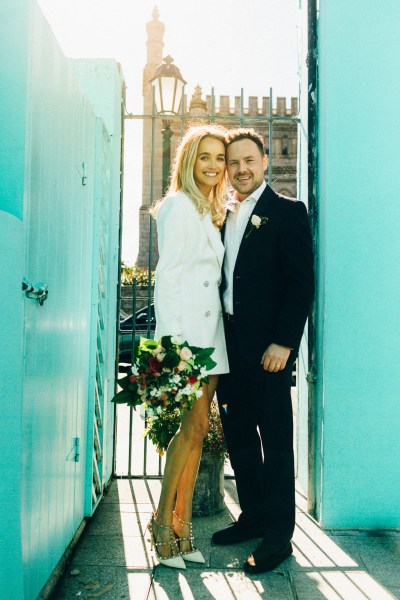 bride and groom stand in between blue wall holding bouquet posing