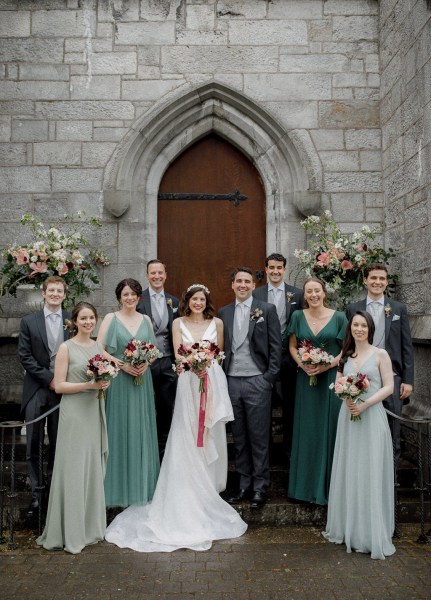 family portrait bride groom in middle exterior church setting with bridesmaids groomsmen and family members either side