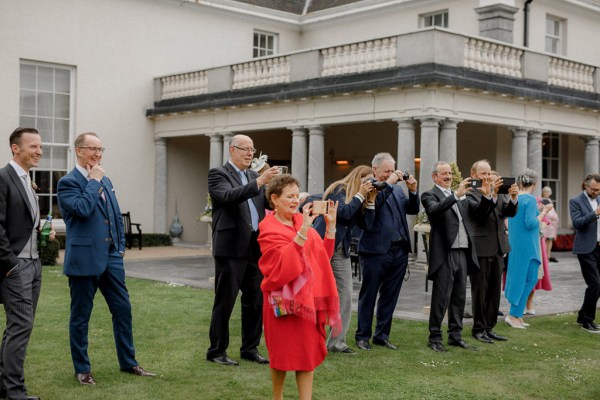 woman in orange red dress gown with guests clapping standing on grass together