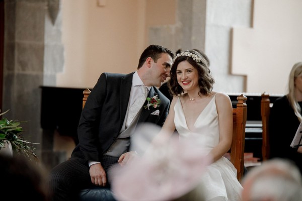 groom whispers into brides ear she smiles as they sit at alter