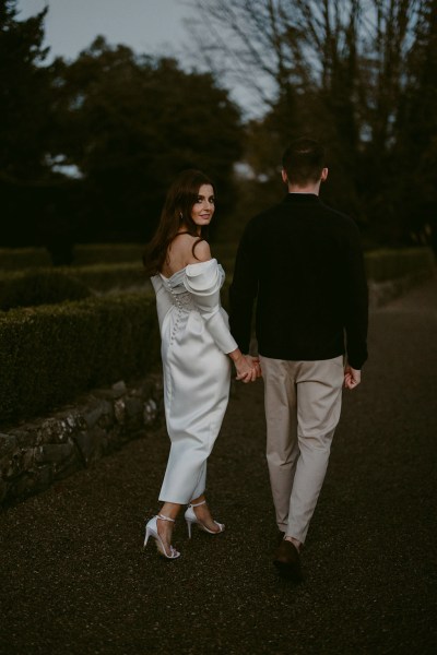 bride looks over her shoulder as she holds hands with groom walking along pathway