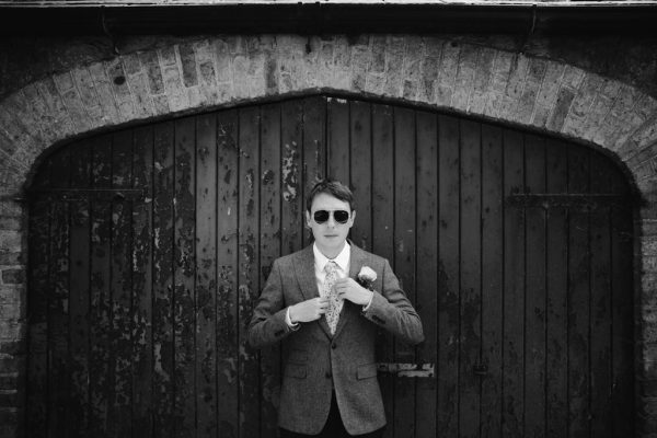 groom looks down in front of wooden gate fixes tie black and white