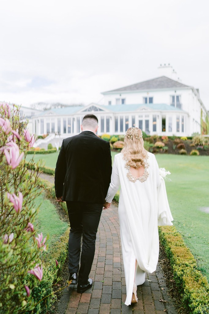 bride and groom walk through the gardens holding hands on pathway walking away from camera