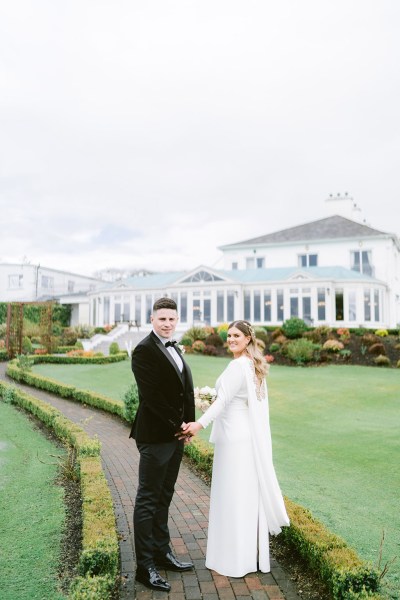 bride and groom walk through the gardens holding hands on pathway over the shoulder