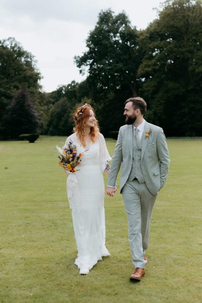 bride and groom walk along the grass together she holds bouquet of flowers