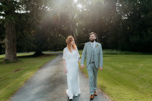 bride and groom walk on the pathway to forest together holding hands she looks at him