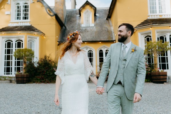 bride and groom walk on the green grass together they look at each other in front of wedding venue holding hands