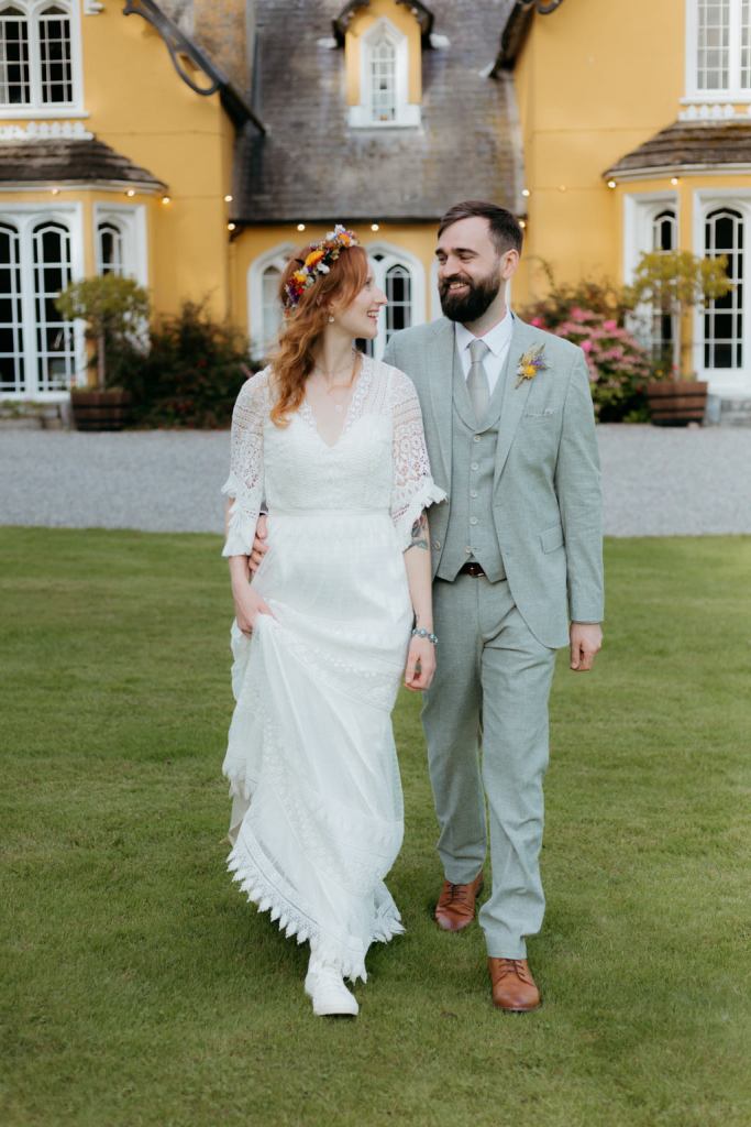 bride and groom walk on the green grass together they look at each other in front of wedding venue