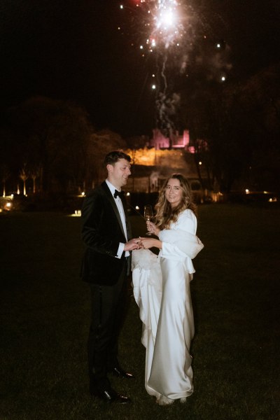 bride and groom face the camera nighttime shot fireworks in background