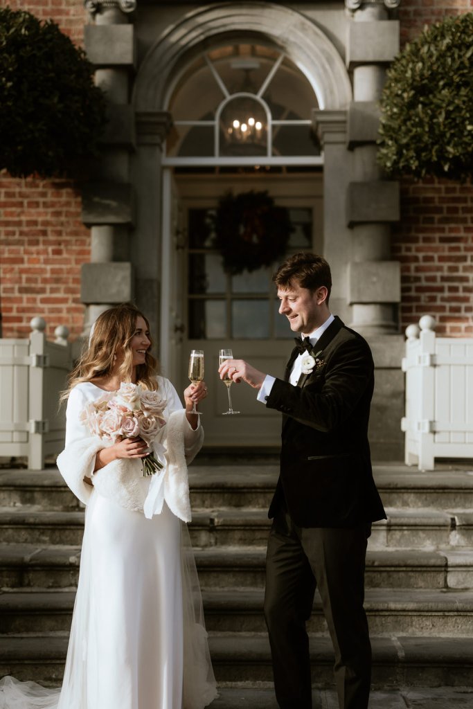 bride and groom cheers with glasses of champagne prosecco in front of steps to wedding venue