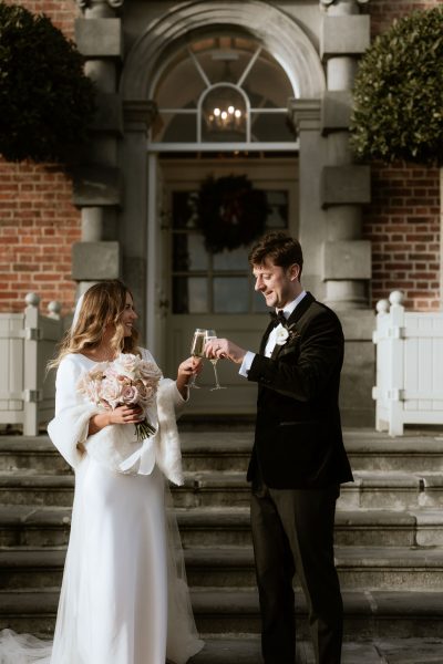 bride and groom cheers with glasses of champagne prosecco in front of steps to wedding venue