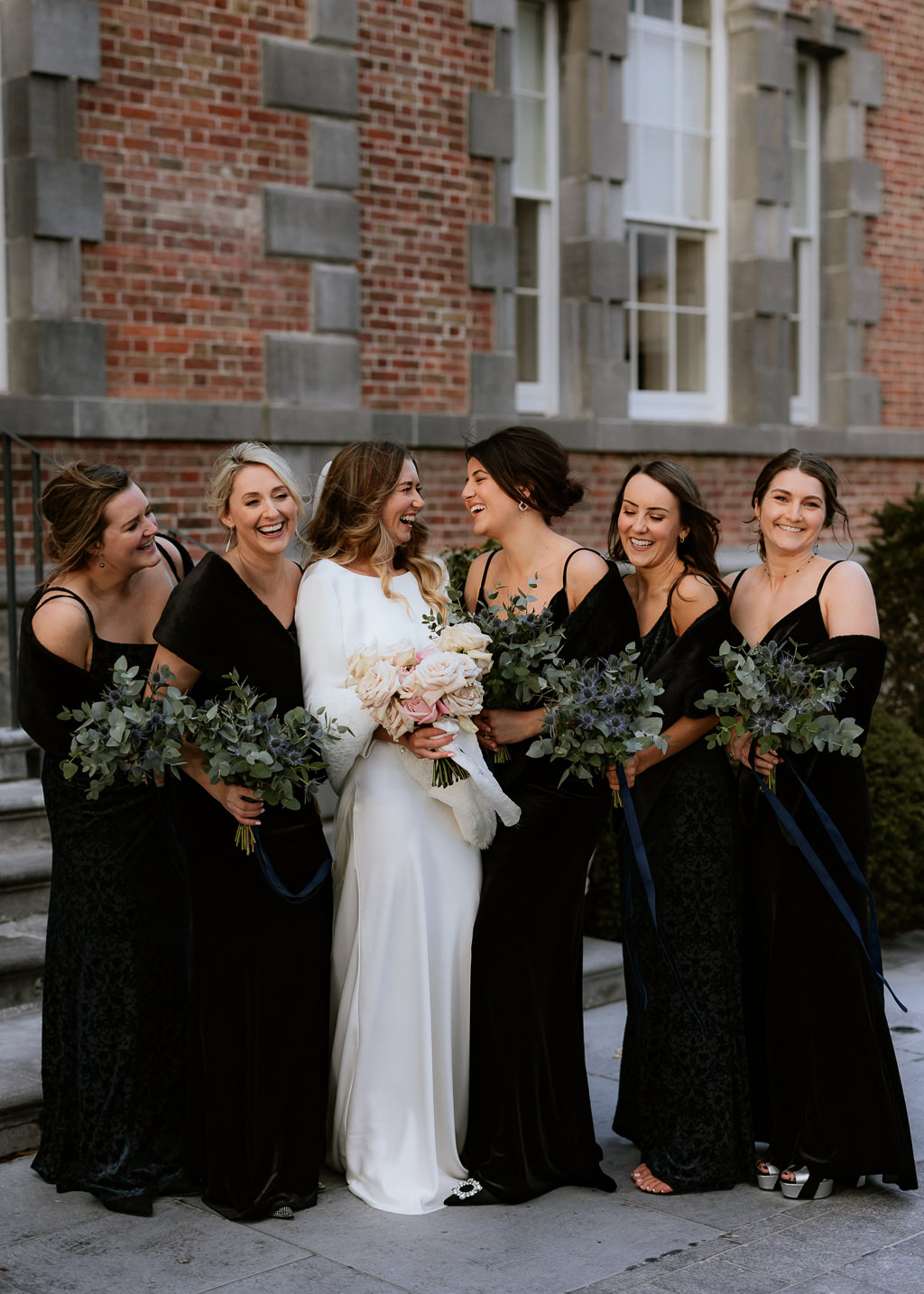 bride and her bridesmaids wearing black dresses holding bouquets smile