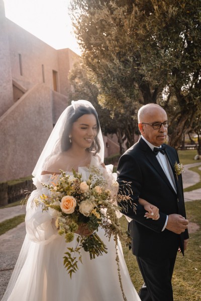 father of the bride walks his daughter towards ceremony she holds bouquet of flowers