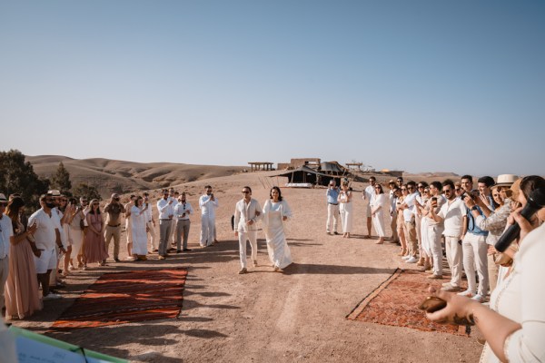 bride and groom walk down sandy pathway surrounded by guests