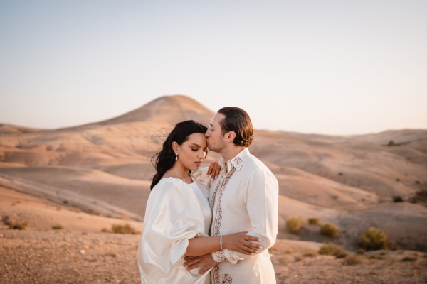 groom kisses top of brides head mountains in background desert