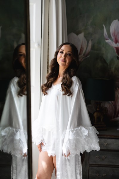 bride stands at window in long white loungewear feather robe