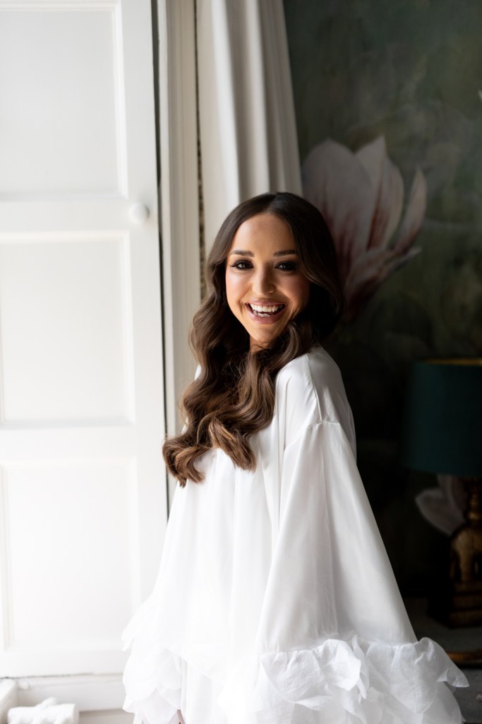 bride stands at window in long white loungewear feather robe