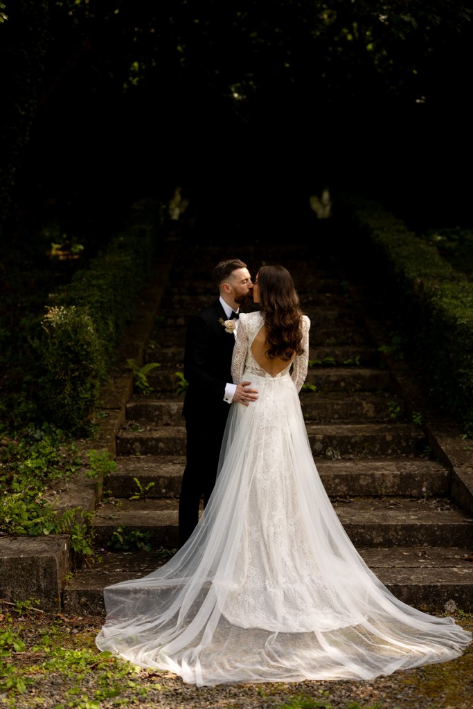 bride shows off back of dress as she stands beside groom steps garden