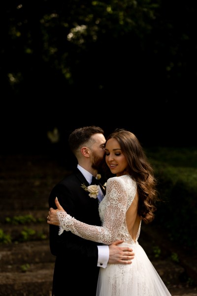 bride looks to the camera groom looks at her they hold each other kiss on the cheek