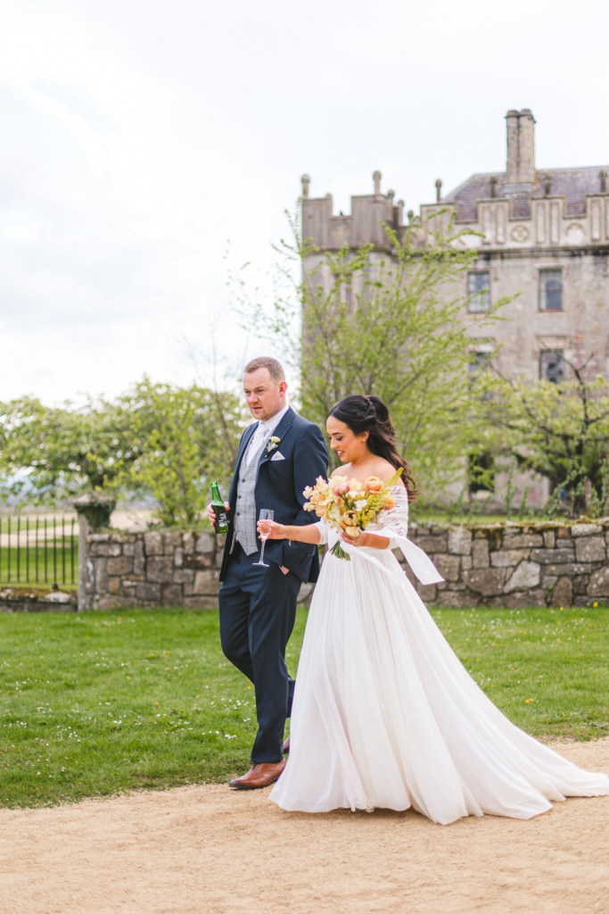 bride and groom walk on the pathway close to grass venue in background she holds bouquet