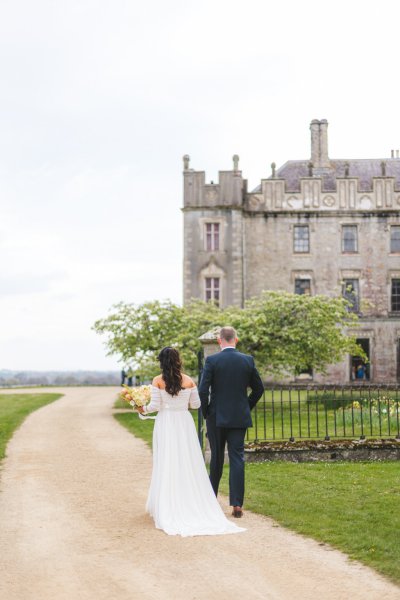 bride and groom walk on the pathway close to grass venue in front she holds bouquet